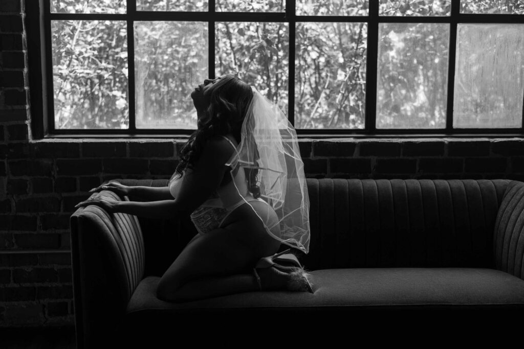 Woman in bridal lingerie, heels, and a wedding veil poses kneeling on a couch for her boudoir photographer