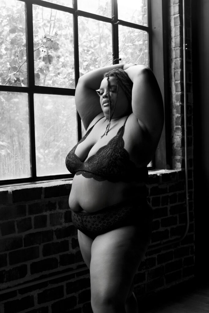 woman standing near a large window with her arms resting above her head, looking down, wearing a 2-piece lingerie outfit during a photo shoot